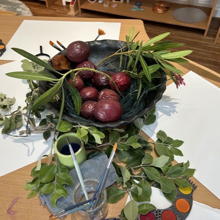 A still-life painting invitation with a bowl of pomegranates, olive branches, and watercolor paints