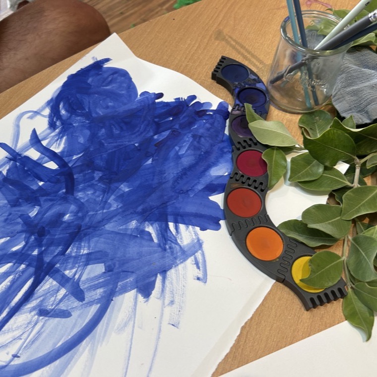A child's blue brush painting beside a watercolor palette and fresh greenery